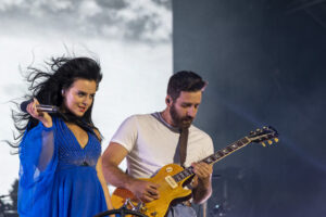 Female singer in a bright blue dress sings into a microphone while a guitarist plays a yellow electric guitar on stage.