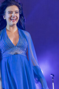 Joyful female singer laughing on stage in a blue, beaded dress under blue-purple lighting