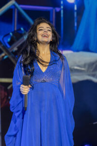Female singer in a bright blue gown, smiling while holding a microphone on stage with blue lighting in the background