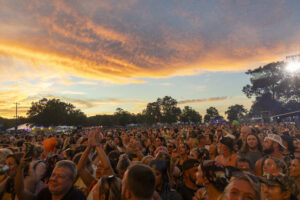 Crowd of festival-goers at an outdoor concert during sunset, with an orange sky and distant stage lighting in the background.