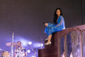 Female singer in a flowing blue dress sits on a balcony edge on stage, holding a microphone and smiling, with a drummer visible in the background and purple stage lights.