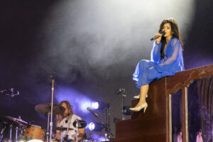 Female singer in a blue flowing dress perched on a piano, microphone in hand, performing on stage with a drummer visible in the background.