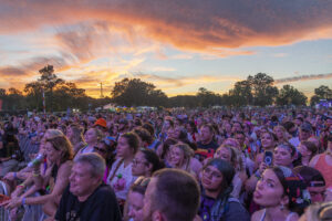 Crowd of festivalgoers packed along a barrier at sunset, facing the stage.