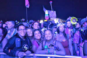 Crowd of smiling festivalgoers pressed against a barrier at night, with purple lighting and glow accessories visible throughout the group.