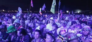 Large nighttime festival crowd with colorful flags, glow accessories, and vibrant outfits.
