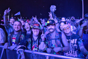 Front-row festival fans behind a barrier, smiling and flashing peace signs under blue stage lights, wearing colorful outfits and accessories.