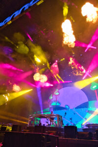 Concert stage with vibrant purple, yellow, and pink lights and large flame effects above a rotating DJ booth. The colorful backdrop features a blue, curved mural and abstract scenery.