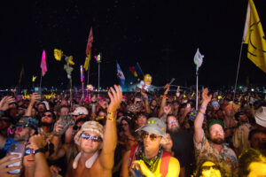 Dense nighttime festival crowd with many raised hands, colorful flags on tall poles, and glow-lit attendees across the scene.