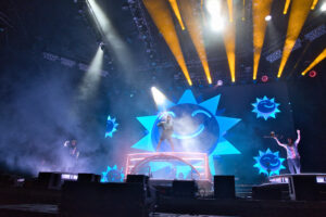 Live concert stage with a singer center stage, blue sunburst screens, and yellow spotlight beams sweeping overhead amid fog.