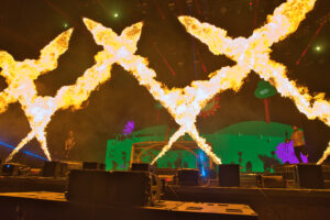 Dramatic stage scene with large flames arcing in zigzag patterns above performers on a green set.