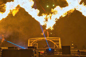 Performer on a platform during a fiery stage show with large orange flames overhead.