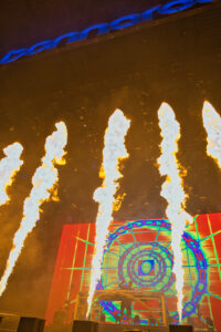 Stage with tall flames shooting upward and a colorful geometric digital backdrop; performers on a raised platform beneath bright lights and stage machinery.
