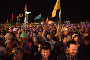 Dense nighttime festival crowd with colorful flags and party gear, packed against a dark sky.