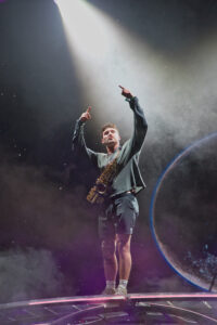 Male saxophonist on a platform, arms raised, under a bright spotlight with a smoky, space-themed stage background.