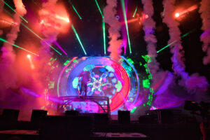 Colorful concert stage with neon lights, smoke plumes, and a large circular LED screen showing abstract visuals; silhouettes of performers on a raised platform in front.
