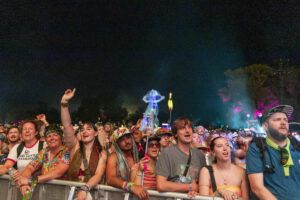 Crowd of festival-goers at night behind a barrier, raised hands, colorful clothing and glow accessories visible at the front.