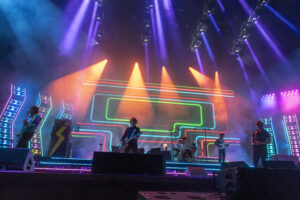 Live concert on a neon-lit stage with guitarists performing amid colorful LED arches and purple-orange beams.
