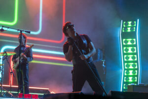 Lead singer with microphone center stage under neon lights, wearing sunglasses and a dark shirt; guitarist in background with a red guitar and colorful backlighting.