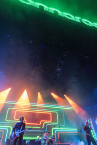 Band performs on a neon-lit stage with green and orange/red light beams and glowing rectangular outlines; guitarist left, drummer center, vocalist right.