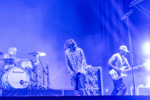 Band on stage performing under blue lights; drummer left, guitarist center, bassist right with microphone stand visible.