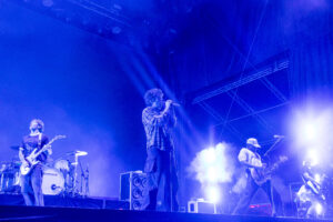 Band performing live on stage with blue lighting; center-stage vocalist with microphone, guitarist on left, drummer in back, foggy atmosphere.