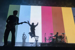 Silhouetted guitarist on stage with a colorful vertical-panel backdrop and a drummer on the right.