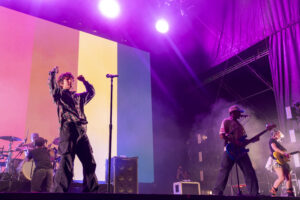 Band performing on stage under pink and purple lights; lead singer raises fists, guitarist and drummer in view, colorful backdrop behind them.