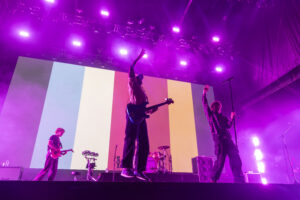 Live band performing on a brightly lit stage with purple lighting and rainbow panels in the background, instruments visible, audience unseen.