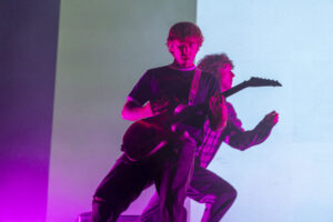 Two guitarists on a lit stage, pink and blue lights; front musician with an electric guitar, eyes closed in performance, another guitarist in the background.