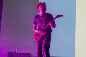 Male guitarist performing on stage under purple-pink lights, playing a pink electric guitar.