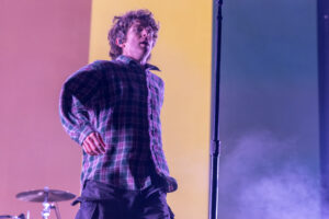 Male musician on stage wearing a plaid shirt, mid-performance under pink and purple lighting, with drum kit visible in the background