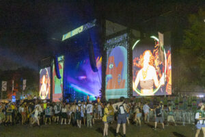 Nighttime outdoor concert with large LED screens displaying performers and a cheering crowd in front of a illuminated stage.