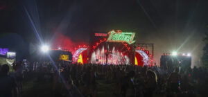 Outdoor night concert with a brightly lit stage, lasers, and a dense crowd; neon sign reads 'The Together'.