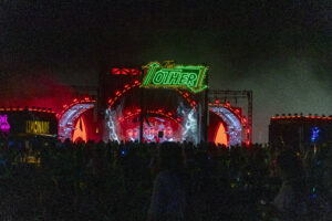 Nighttime outdoor concert with a neon green 'The Other' sign above a large, illuminated stage and cheering crowd.