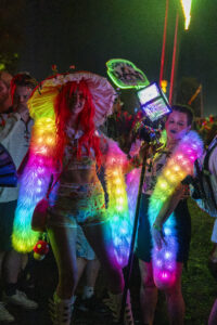 People at a nighttime festival wearing colorful neon outfits and fluffy rainbow sleeves under bright lights.