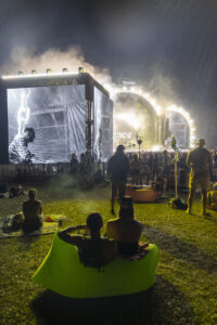 Nighttime outdoor concert with a large stage, circular light arch, big screen, and a crowd on grass; a person sits in a bright green inflatable chair in the foreground.