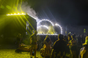 Crowd at a nighttime outdoor music festival with bright yellow stage lights and a circular lit stage backdrop behind the performers on a dark sky.