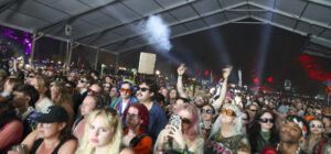 Large crowd at a nighttime outdoor concert under a white canopy, with smoke and colorful lights in the background and hands raised.