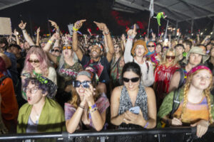 Large crowd at a nighttime concert, many people with arms raised, colorful outfits and accessories, behind a barrier on a lively festival stage.