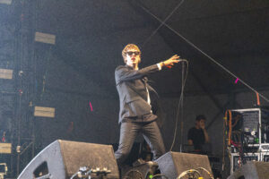 Male singer in a black suit and sunglasses performing on stage with mic stand, stage lighting and equipment around him