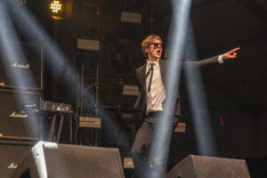 DJ in a suit and sunglasses points toward the crowd on a dark stage with Marshall amps and bright beams of light nearby.