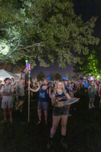 Nighttime festival crowd under a leafy tree canopy; woman with tattoos in the foreground holds a drum.