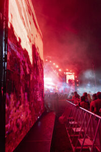 Large crowd at a nighttime concert with red stage lighting and a towering LED screen on the left.