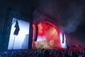 Concert crowd facing a large stage with red laser lights and towering metal scaffolding in the background, at night or dusk.