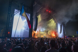 Crowd at a nighttime outdoor concert with a large stage, lighting, and a waving flag in the foreground.