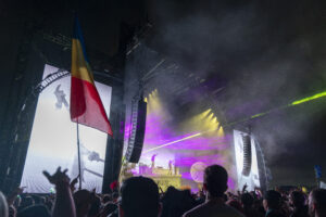 Romanian flag waving in front of a large illuminated concert stage with colorful lights and a foggy night backdrop, crowd cheering in the foreground.