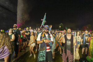 Large nighttime festival crowd wearing colorful outfits and glow accessories, with a tall teal and pink inflatable sculpture in the background.
