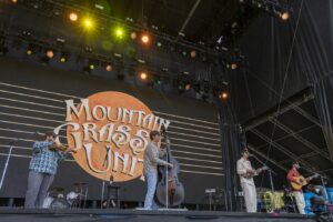 Band with four musicians performing on an outdoor stage; a large screen behind shows an orange circle and the words 'Mountain Grass Inc'. The lineup includes a violinist, a stand-up bassist, a mandolin player, and a guitarist/singer.