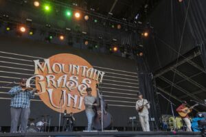 Live bluegrass band performing on an outdoor stage with a large screen backdrop featuring an orange circle and stylized text behind them, under colorful stage lights.