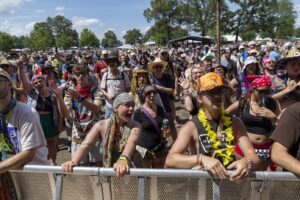 Crowd at an outdoor festival behind a metal barricade, wearing colorful clothes and accessories on a sunny day.